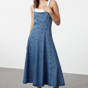 K&ouml;nnte beinhalten: Ein blaues Jeans-Midikleid mit einem figurbetonten Oberteil und einem ausgestellten Rock. Das Kleid hat einen quadratischen Ausschnitt und d&uuml;nne Tr&auml;ger.