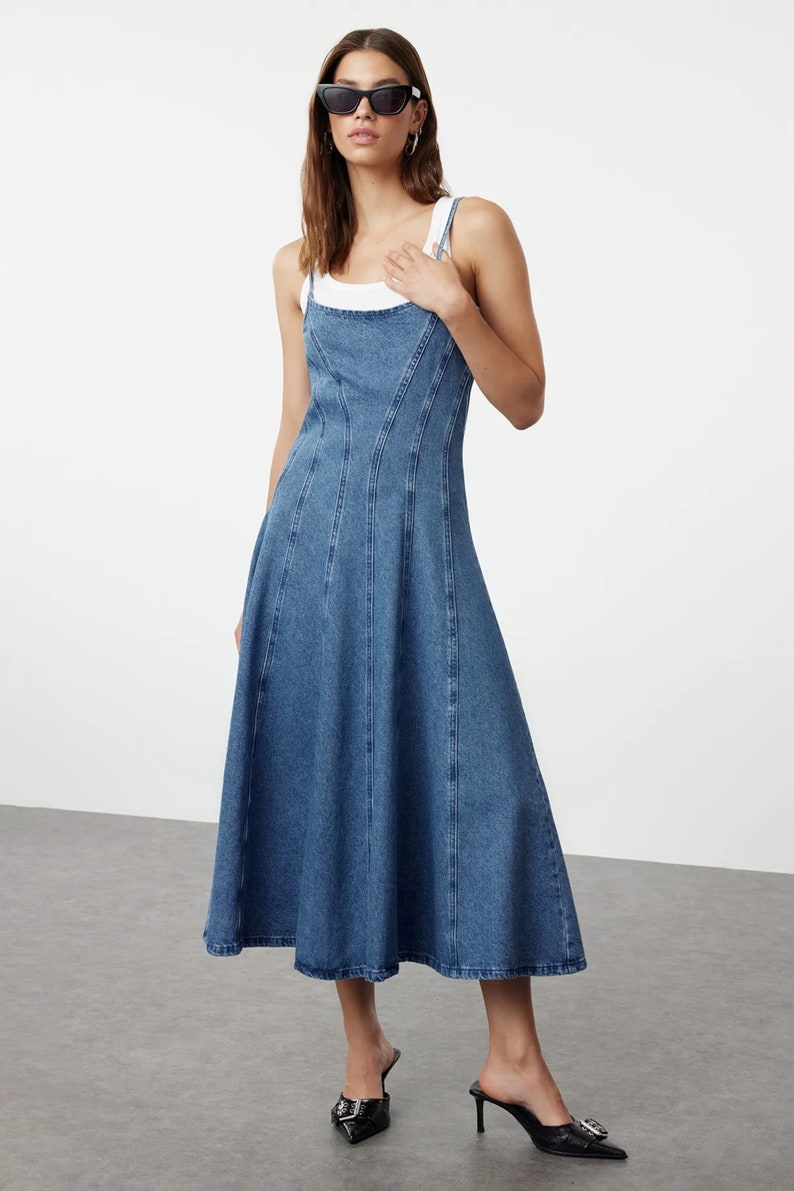 K&ouml;nnte beinhalten: Ein blaues Jeans-Midikleid mit einem figurbetonten Oberteil und einem ausgestellten Rock. Das Kleid hat einen quadratischen Ausschnitt und d&uuml;nne Tr&auml;ger.