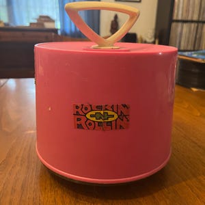 May include: A bright pink, cylindrical record case with a cream-colored handle. The case features a sticker with the words "Rockin' Rollin'" in a retro font. The case is sitting on a wooden surface.