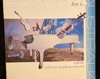 May include: A white grand piano with a red bird perched on top. Three violins are floating in the air above the piano. The text "Elton John live in Australia with the Melbourne Symphony Orchestra" is printed on the album cover.