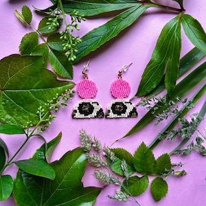 Könnte beinhalten: Handgefertigte Perlenohrringe mit einem rosa Oberteil und einem schwarz-weiß gemusterten Unterteil. Die Ohrringe sind vor einem lila Hintergrund mit grünen Blättern und Laub abgebildet.