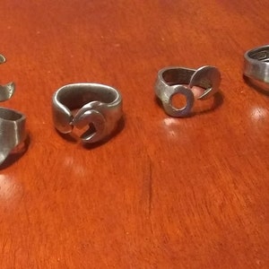 May include: Five silver rings made from repurposed wrenches. The rings are arranged in a row on a wooden surface.