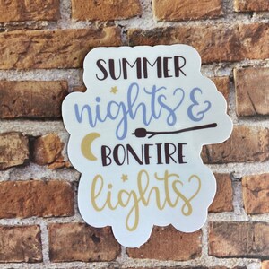 May include: A white sticker with the words "SUMMER nights & BONFIRE lights" in various fonts and colors. The design includes a crescent moon, stars, and a marshmallow on a stick. The sticker is on a brick background.