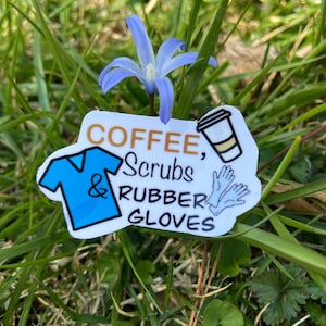 May include: A sticker with the words "COFFEE, Scrubs & RUBBER GLOVES" and illustrations of a coffee cup, scrubs, and gloves. A small blue flower is in the background, with green grass.