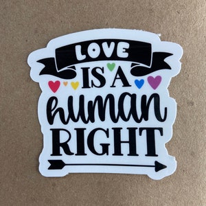 May include: White sticker with the text "LOVE IS A HUMAN RIGHT" in black, with colorful hearts and an arrow. The word "LOVE" is on a black banner. The sticker is on a brown surface.
