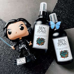 May include: A black bottle of "Ben Solo Hero Edition" body and room linen fragrance mist, with a "Star Wars" figurine and a blue butterfly. The bottle has a white label with text and a decorative design. The figurine is of a character holding a lightsaber.