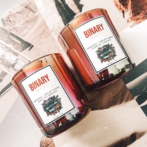May include: Two amber glass candle jars with iridescent shimmer. Each jar has a white label with the text "BINARY Sungel" and "WOOD WICK 100% SOY CANDLE ALMOND SALT SANDALWOOD" and a floral design. The jars are sitting on a brown and white background.