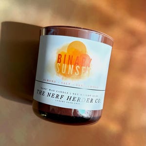 May include: A brown glass jar candle with a white label featuring a watercolor sunset graphic. The text reads Binary Sunset, Almond Salt Sandalwood, Sand Wax Candle, Net Weight 13 oz, and The Nerf Herder Co. Luxury Geek Chic in black font.