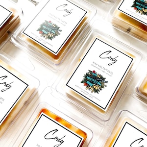 May include: A set of six wax melts in clear plastic containers. Each container has a label with the name "Cody" and the text "Wax Melts/Tarts Cedarwood Grey Vetiver" with a floral design. The wax melts are a light amber colour.