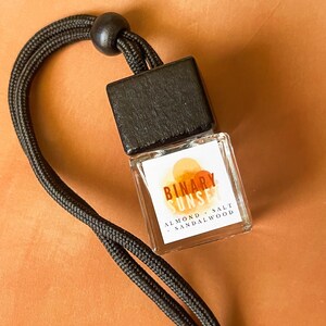 Op de afbeelding: Een kleine, heldere glazen fles met een zwarte houten dop en een zwart koord. Het etiket luidt "BINARY SUNSET" met "ALMOND - SALT - SANDALWOOD" eronder. De achtergrond is effen oranje-bruin.