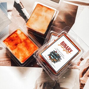 May include: Three wax melts in clear plastic containers. The wax melts are orange and brown. The container on the right has a label that reads "BINARY sunset WAX MELTS/TARTS ALMOND SALT & SANDALWOOD NET WEIGHT 2.5 OZ BLEND OF 100% NATURAL SOY AND BEESWAX".
