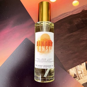 May include: A clear glass bottle with a gold cap and a white label. The label says "BINARY SUNSET BODY ROOM LINEN FRAGRANCE MIST NET WEIGHT 2.5 FL/100 ML THE NERF HERDER CO. LUXURY GEEK CHIC".