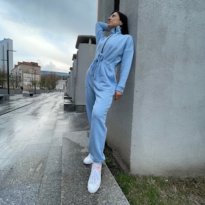 May include: A light blue jumpsuit with a zipper and a belt. The jumpsuit has long sleeves and a high neck. The person is wearing white sneakers.