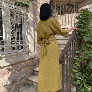 Pu&ograve; includere: Un lungo abito di lino verde oliva con cintura a nodo. L'abito &egrave; indossato da una persona in piedi su una serie di gradini in pietra.
