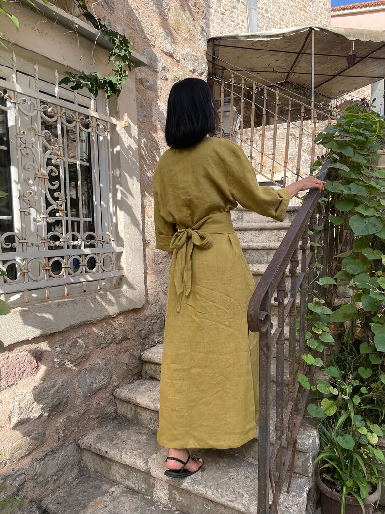 Pu&ograve; includere: Un lungo abito di lino verde oliva con cintura a nodo. L'abito &egrave; indossato da una persona in piedi su una serie di gradini in pietra.