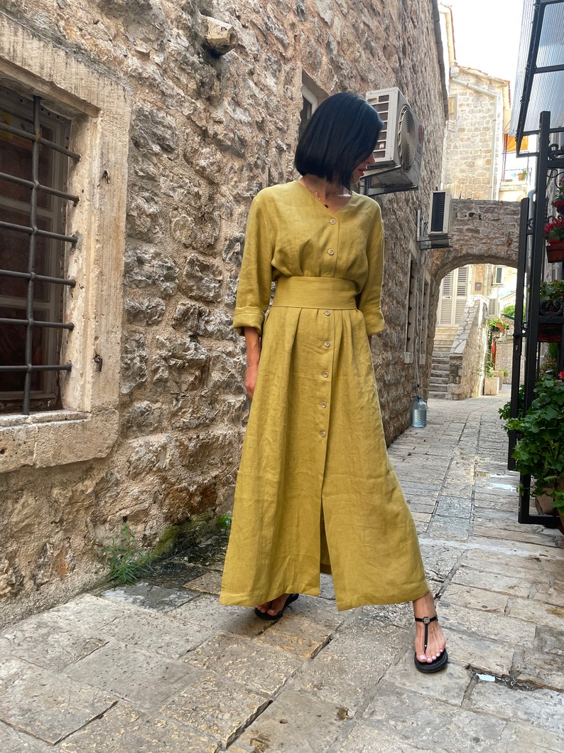 Pu&ograve; includere: Un lungo abito in lino giallo senape con bottoni sul davanti e una cintura. L'abito ha una vestibilit&agrave; rilassata e una gonna fluida.