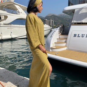 Pu&ograve; includere: Una donna indossa un abito maxi in lino giallo senape con uno spacco laterale e una fascia abbinata, si trova su un molo vicino a uno yacht bianco con la scritta "BLU" sul lato.