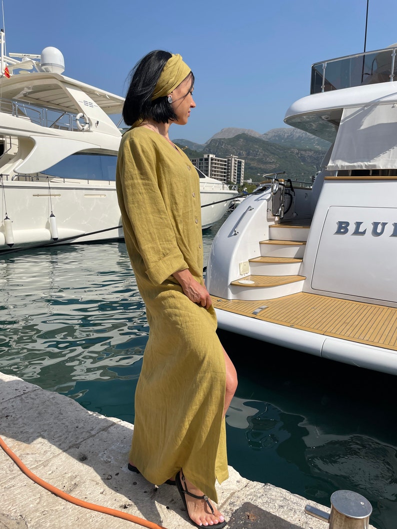 Pu&ograve; includere: Una donna indossa un abito maxi in lino giallo senape con uno spacco laterale e una fascia abbinata, si trova su un molo vicino a uno yacht bianco con la scritta "BLU" sul lato.