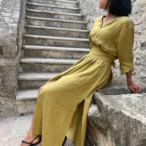 Pu&ograve; includere: Una donna indossa un lungo abito di lino giallo senape con uno spacco laterale e una cintura abbinata. &Egrave; seduta su un bordo di pietra con le gambe incrociate e indossa sandali neri.