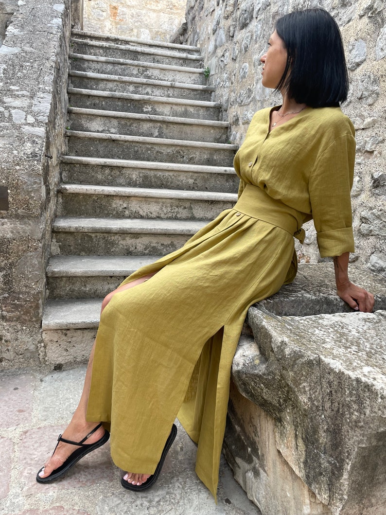 Pu&ograve; includere: Una donna indossa un lungo abito di lino giallo senape con uno spacco laterale e una cintura abbinata. &Egrave; seduta su un bordo di pietra con le gambe incrociate e indossa sandali neri.