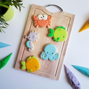 Puede incluir: Cinco galletas de azúcar decoradas en una tabla de cortar de madera. Las galletas tienen forma de cangrejo, caballito de mar, pulpo y dos peces. Las galletas están decoradas con glaseado de colores y tienen caras lindas.