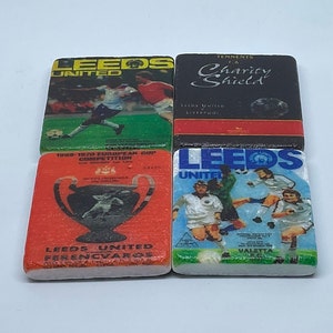 May include: Four square coasters with Leeds United football designs. One shows a match, another the Charity Shield logo, a third the European Cup, and the last players in action. Each coaster has a different design, with text and imagery.