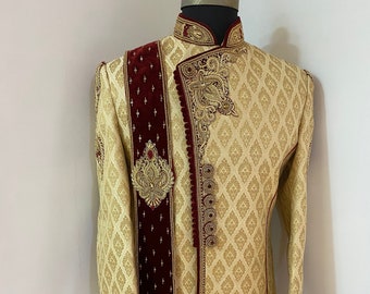 May include: A gold and cream patterned sherwani with a maroon velvet shawl. The sherwani has gold embroidery and embellishments.