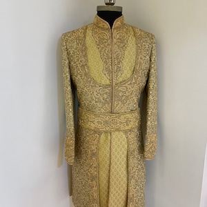 May include: A gold-colored, long-sleeved jacket with intricate embroidery and a mandarin collar. The jacket features a matching embroidered belt and a pleated lower section. The garment is displayed on a mannequin.