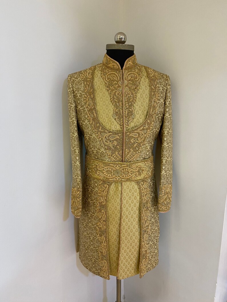 May include: A gold-colored, long-sleeved jacket with intricate embroidery and a mandarin collar. The jacket features a matching embroidered belt and a pleated lower section. The garment is displayed on a mannequin.