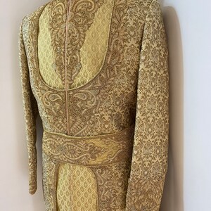 May include: A gold-colored, intricately embroidered sherwani suit displayed on a mannequin. The formal wear features a high collar, ornate patterns, and a decorative belt. The fabric has a subtle sheen, and the design includes detailed floral and paisley motifs.