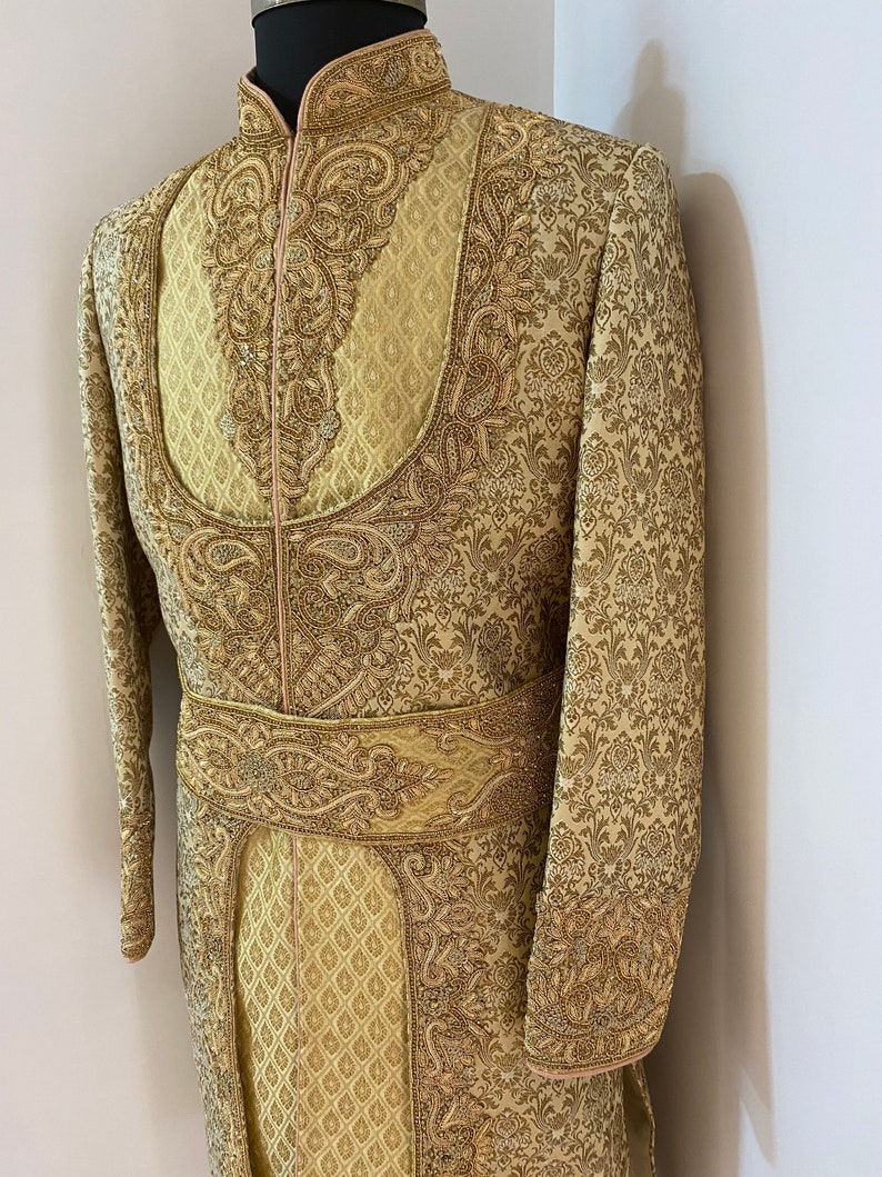 May include: A gold-colored, intricately embroidered sherwani suit displayed on a mannequin. The formal wear features a high collar, ornate patterns, and a decorative belt. The fabric has a subtle sheen, and the design includes detailed floral and paisley motifs.