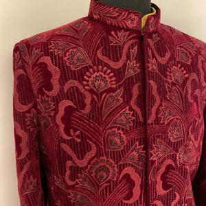 May include: A maroon velvet jacket with a mandarin collar and a floral pattern. The jacket has a button closure and long sleeves.