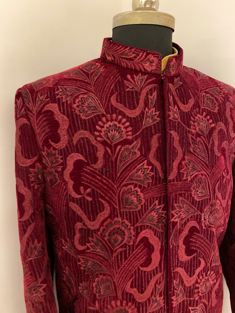 May include: A maroon velvet jacket with a mandarin collar and a floral pattern. The jacket has a button closure and long sleeves.