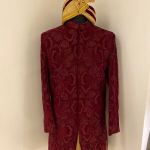 May include: A maroon velvet kurta with a floral pattern and a gold and maroon turban. The kurta has a long, flowing design and a stand-up collar.