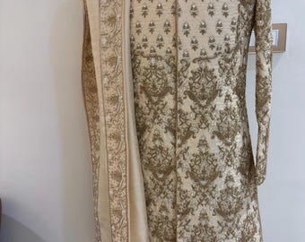 May include: A cream-coloured embroidered men's kurta with a matching dupatta. The kurta has a mandarin collar and a long, flowing design. The embroidery is intricate and features gold thread and beads.