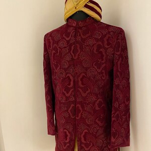 May include: A maroon velvet kurta with a floral pattern and a gold embroidered trim. The kurta has a mandarin collar and long sleeves. A gold turban with a red band is on top of the kurta.