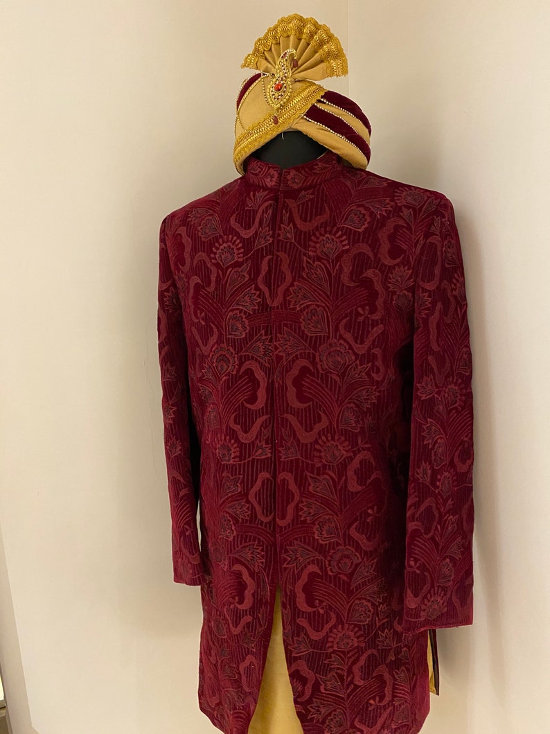 May include: A maroon velvet kurta with a floral pattern and a gold embroidered trim. The kurta has a mandarin collar and long sleeves. A gold turban with a red band is on top of the kurta.