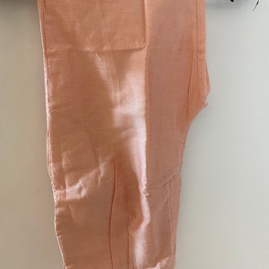 May include: Peach-colored pants hanging on a black hanger. The pants are made of a shiny, silky material. The pants are folded and appear to be new. The pants are a solid color and have a simple design. The pants are likely part of a formal outfit.