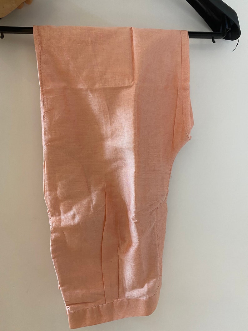 May include: Peach-colored pants hanging on a black hanger. The pants are made of a shiny, silky material. The pants are folded and appear to be new. The pants are a solid color and have a simple design. The pants are likely part of a formal outfit.