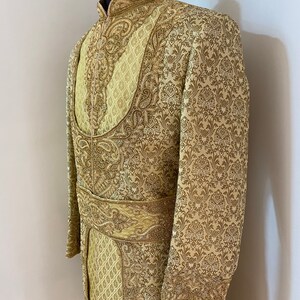 May include: A gold-colored, ornate sherwani suit with intricate embroidery and a mandarin collar. The suit features detailed patterns and a matching belt, displayed on a mannequin. The fabric has a subtle sheen, enhancing the luxurious appearance.