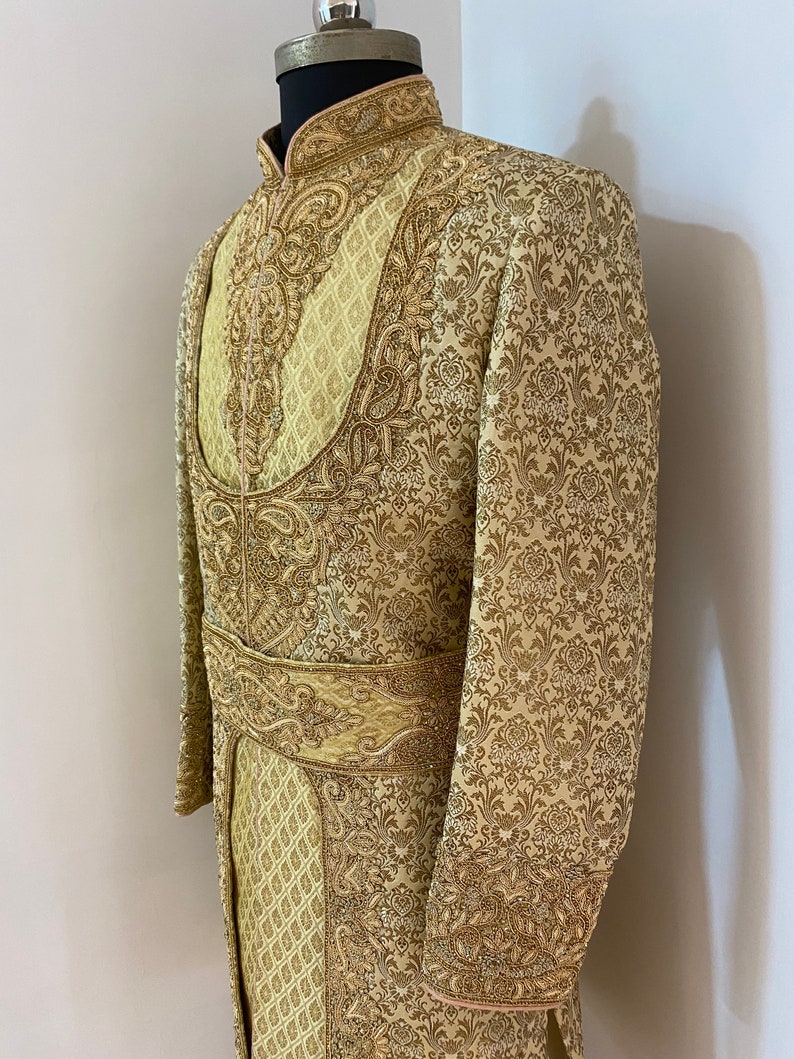 May include: A gold-colored, ornate sherwani suit with intricate embroidery and a mandarin collar. The suit features detailed patterns and a matching belt, displayed on a mannequin. The fabric has a subtle sheen, enhancing the luxurious appearance.