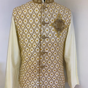 May include: A white waistcoat with a gold and white geometric pattern and a gold beaded embellishment. The waistcoat has a mandarin collar and a button closure.