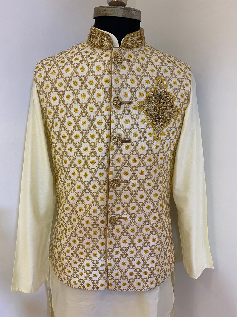 May include: A white waistcoat with a gold and white geometric pattern and a gold beaded embellishment. The waistcoat has a mandarin collar and a button closure.