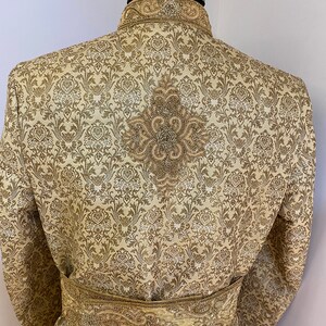 May include: A gold-colored, patterned jacket with a mandarin collar and ornate embroidery. The back of the jacket features a detailed floral design. A matching gold belt with intricate detailing is wrapped around the waist. The jacket is displayed on a mannequin.