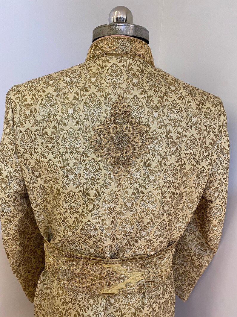 May include: A gold-colored, patterned jacket with a mandarin collar and ornate embroidery. The back of the jacket features a detailed floral design. A matching gold belt with intricate detailing is wrapped around the waist. The jacket is displayed on a mannequin.