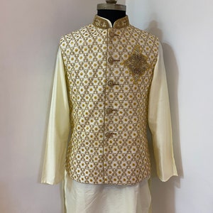 May include: A white kurta with a gold embroidered waistcoat. The waistcoat has a geometric pattern and a gold embellishment on the front.