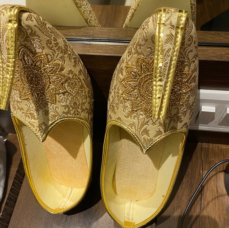 May include: A pair of ornate, gold-colored slip-on shoes with intricate floral embroidery. The shoes feature a pointed toe and a decorative gold trim along the edges. The design includes a central beaded floral pattern.