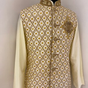 May include: A white waistcoat with a gold embroidered pattern and a gold beaded embellishment on the front. The waistcoat has a mandarin collar and a button closure.