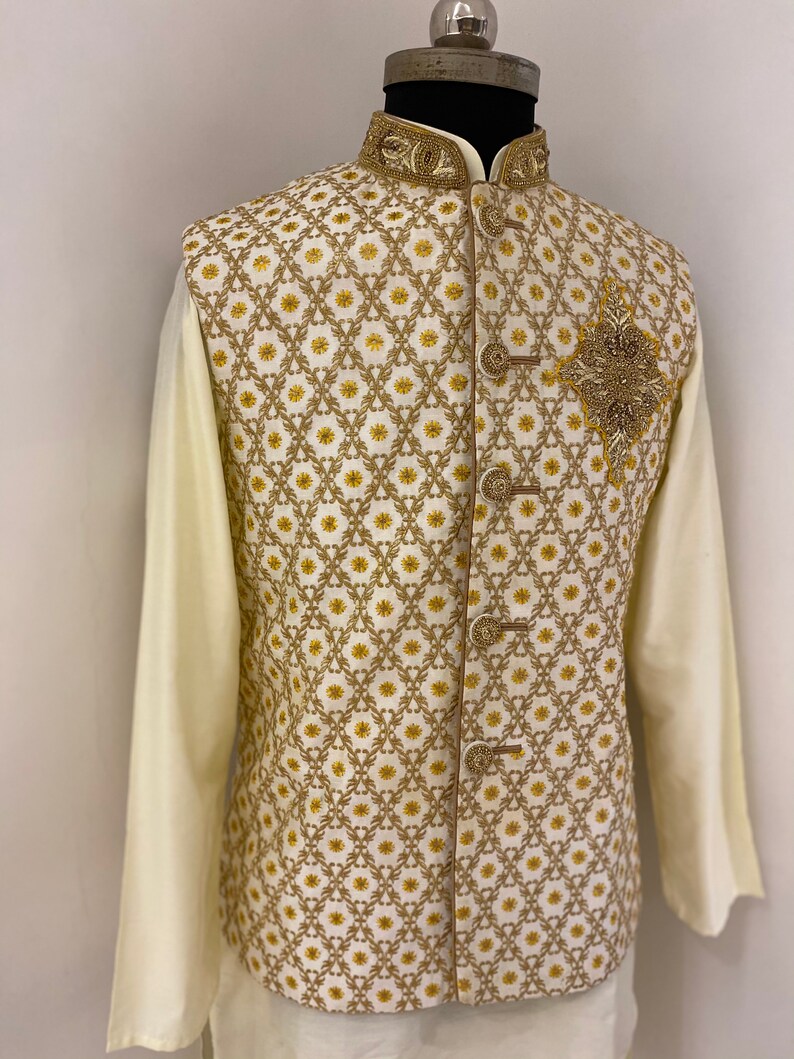 May include: A white waistcoat with a gold embroidered pattern and a gold beaded embellishment on the front. The waistcoat has a mandarin collar and a button closure.