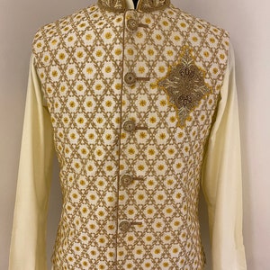 May include: A white waistcoat with a gold and yellow geometric pattern and a gold embroidered design on the front. The waistcoat has a mandarin collar and a button closure.